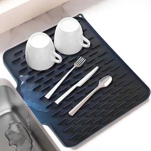 Dark silicone dish drying mat with water drainage, holding two cups and cutlery next to a sink