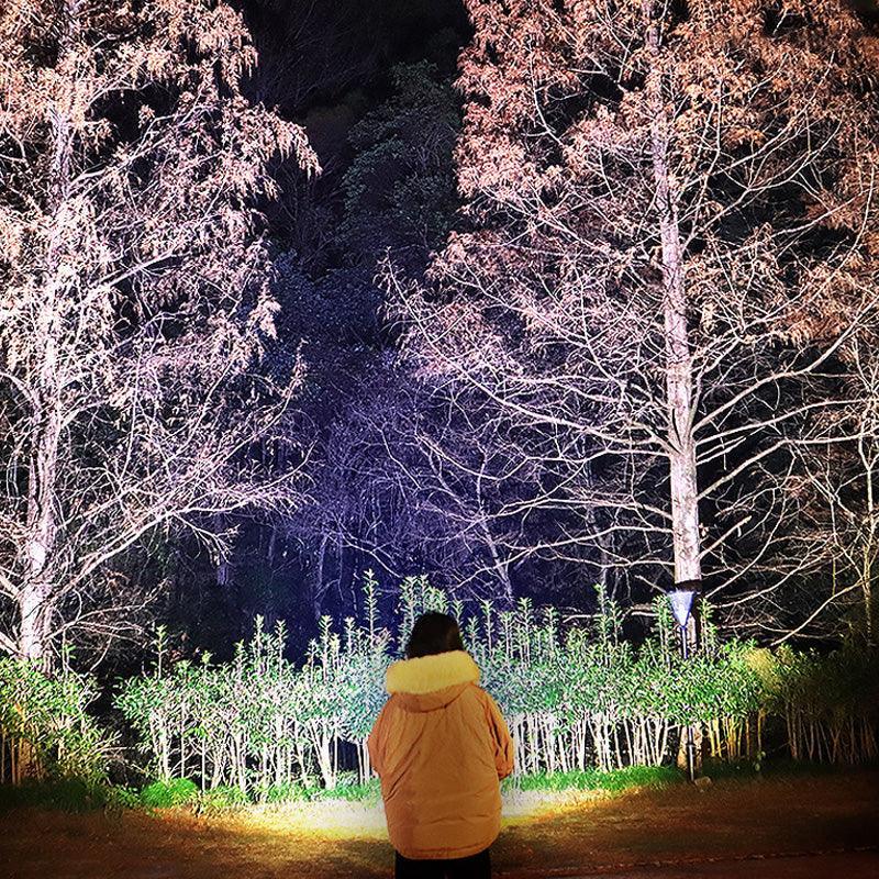 Person in beige winter coat standing outdoors at night with illuminated trees and bushes