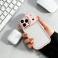 Hand holding iPhone with pink case near wireless earbuds, keyboard, and mouse on desk