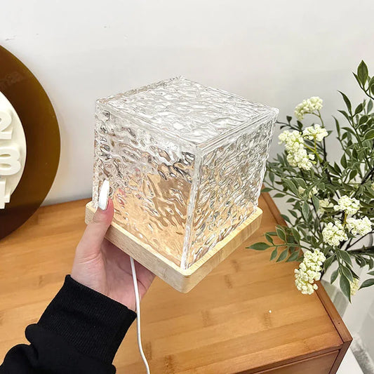 Hand holding a modern glass cube table lamp with wooden base on a wooden desk, home decor by clarioy