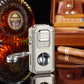 Premium metal cigar lighter and cutter on reflective surface with cigar, leather case, and liquor in background