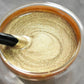 Close-up of gold glitter paint or gel in a clear jar, with a black spatula stirring, clarioy homeware.